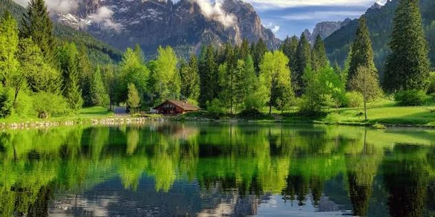 Lago con reflejos de árboles, una pequeña construcción de madera junto a la orilla y montañas al fondo. Imagen aportada por Pablo Gustavo Leonardi como paisaje del entorno de Primiero; autor y localización precisa pendientes de confirmar.